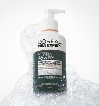 Flacon blanc de nettoyant purifiant L'Oréal Men Expert Derma Power avec pompe, entouré de bulles de savon blanches sur un fond gris.