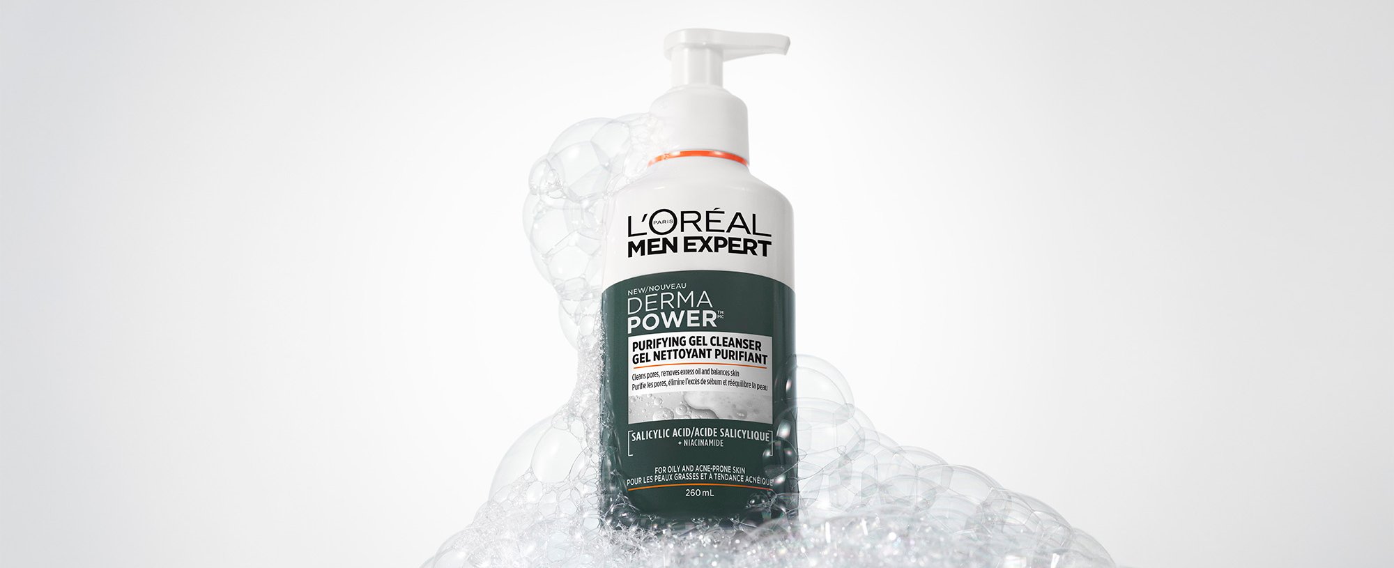 White bottle of L'Oréal Men Expert Derma Power Purifying Gel Cleanser with a pump, surrounded by white soap bubbles on a grey background.