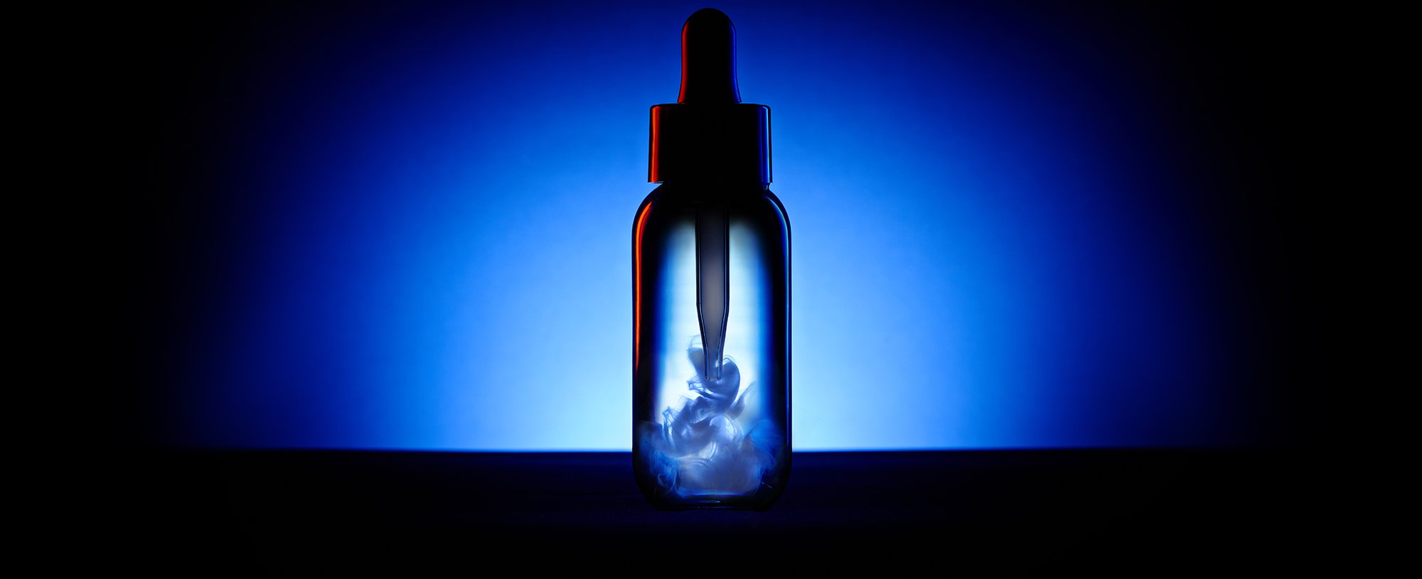 Flacon sombre avec pipette contenant un liquide blanc en suspension, sur un fond bleu et noir profond.