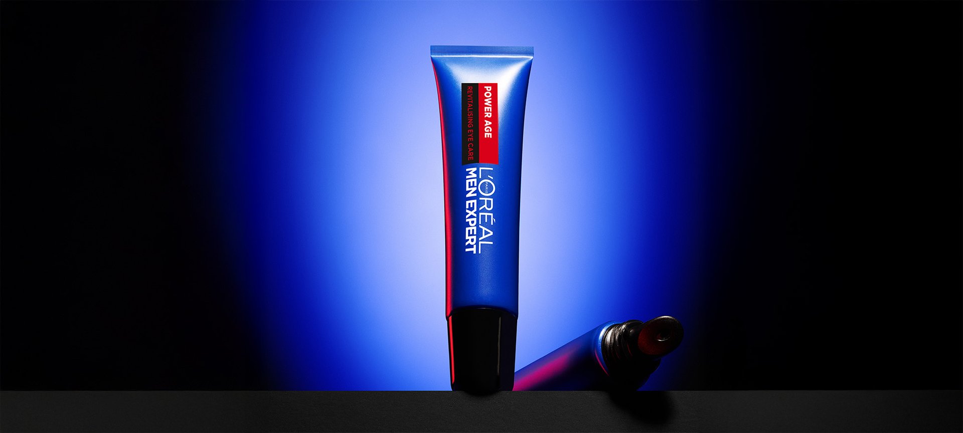 Blue tube of L'Oréal Men Expert Power Age Revitalising Eye Care lying on a dark surface, with a blue background.