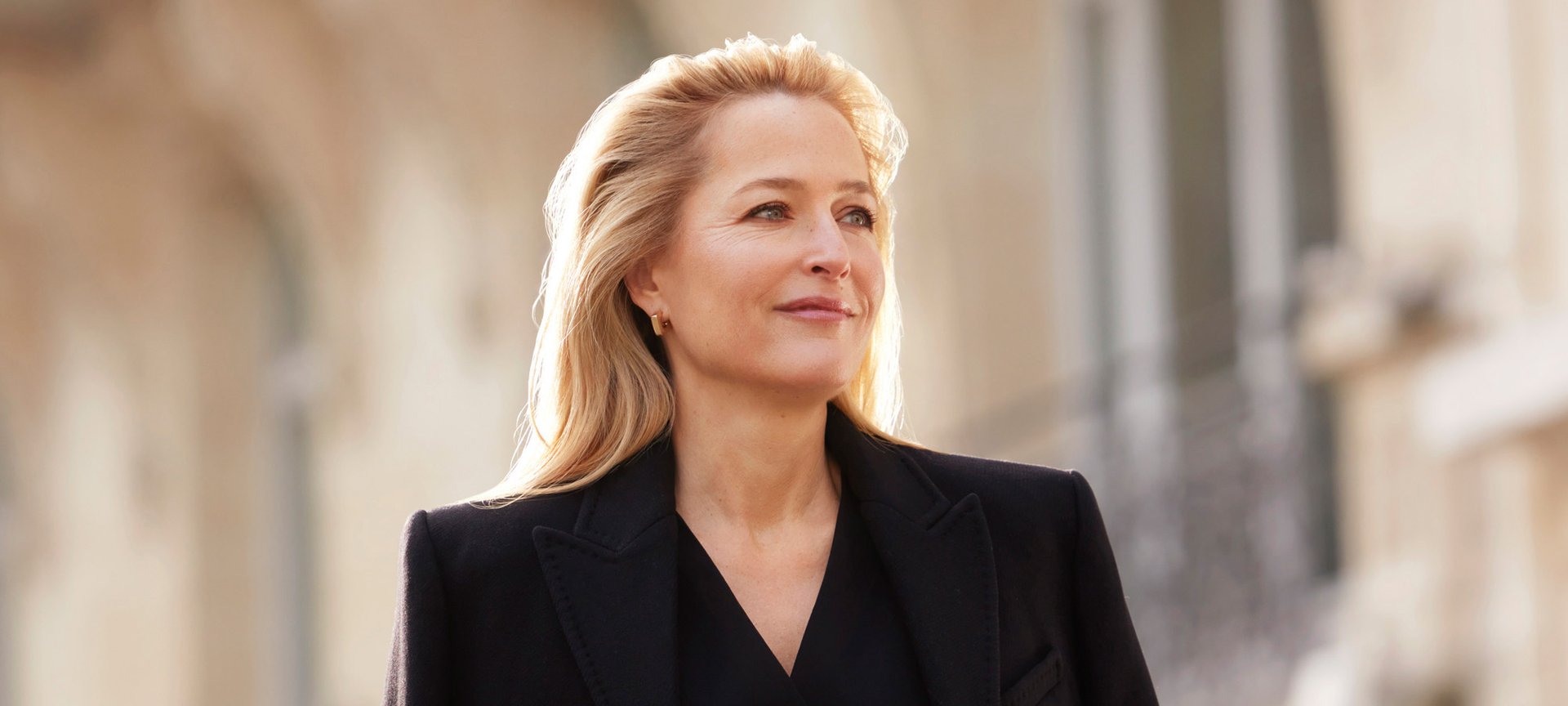 Portrait de Gillian Anderson, souriante et regardant au loin, les cheveux blonds, portant une veste sombre, avec un arrière-plan flou et ensoleillé