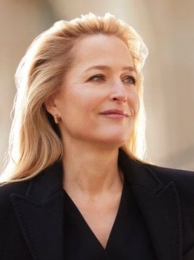 Portrait de Gillian Anderson, souriante et regardant au loin, les cheveux blonds, portant une veste sombre, avec un arrière-plan flou et ensoleillé