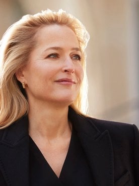 Portrait of Gillian Anderson, smiling and looking into the distance, with blonde hair, wearing a dark jacket, against a soft, sunlit background