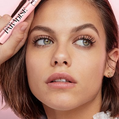 A woman with short brown hair holds a pink L'Oréal Lash Paradise mascara, looking upwards to reveal her beautifully fanned and voluminous eyelashes.