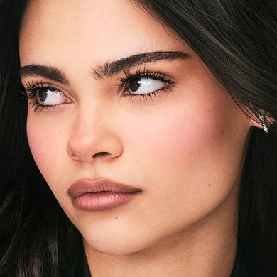 A woman with dark hair and striking eyes looks subtly to the side, showcasing her naturally enhanced eyelashes with mascara, illustrating definition and beauty.