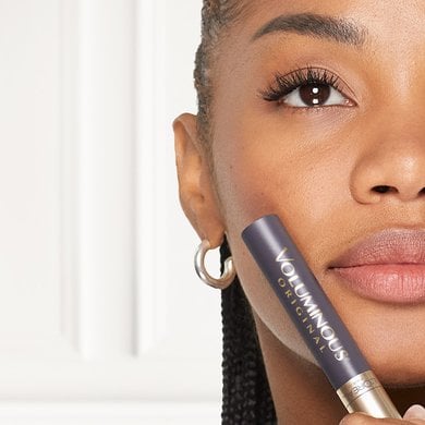 Une jeune femme souriante tient un mascara L'Oréal Voluminous Original, montrant ses cils longs et volumineux après application, soulignant l'effet volumisant du produit.