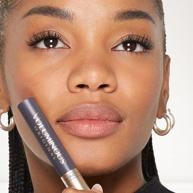 A smiling young woman holds a L'Oréal Voluminous Original mascara, showcasing her long, full eyelashes after application, emphasizing the product's volumizing effect.
