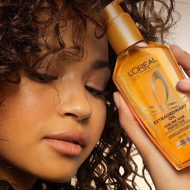 Close-up of a woman with very dark curly hair, eyes closed, holding a golden bottle of L'Oréal 'Sublime Hair Perfector' Extraordinary Hair Oil near her face