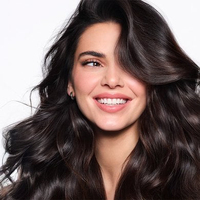 Gros plan sur Kendall Jenner souriant largement, montrant ses dents, avec des cheveux bruns foncés et très volumineux, coiffés en vagues. Elle regarde vers le côté droit de l'image