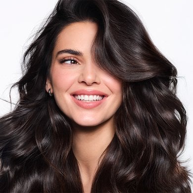 Close-up of Kendall Jenner smiling broadly, showing her teeth, with very voluminous dark brown hair styled in waves. She is looking towards the right side of the image