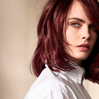 Portrait of Cara Delevingne, looking over her right shoulder, with short to medium-length auburn or burgundy hair styled in soft waves. She is wearing a white shirt and her blue eyes are prominent