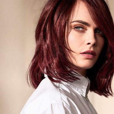 Portrait de Cara Delevingne, regardant par-dessus son épaule droite, avec des cheveux courts à mi-longs auburn ou bordeaux coiffés en vagues souples. Elle porte une chemise blanche et ses yeux bleus sont mis en valeur
