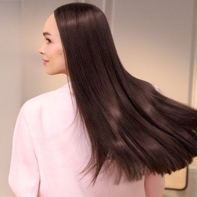Une femme de dos, ses longs cheveux bruns très raides et lisses flottent légèrement derrière elle. Elle porte une chemise rose pâle et regarde vers la droite