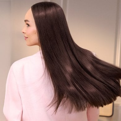 A woman from behind, her long, very straight and sleek brown hair slightly flowing behind her. She is wearing a light pink shirt and looking to her right