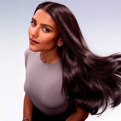 Une femme aux longs cheveux bruns foncés et brillants en mouvement, regardant l'objectif. Elle porte un haut gris et a du rouge à lèvres foncé
