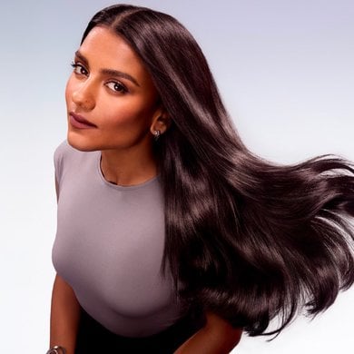 A woman with long, dark, shiny hair in motion, looking at the camera. She is wearing a grey top and has dark lipstick