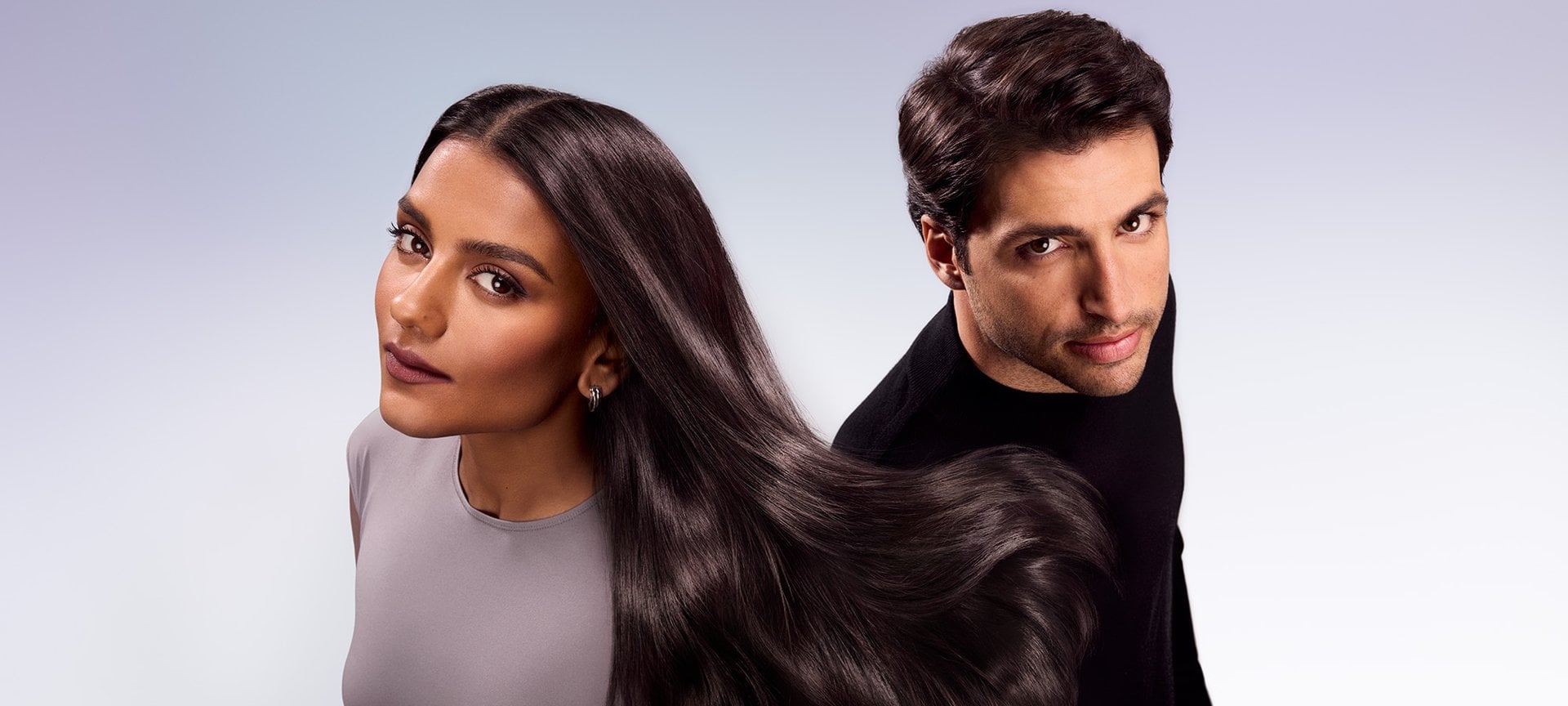 A man and a woman standing side by side, the man looking at the camera while the woman looks over her shoulder. Their dark brown hair is long and shiny, with the woman's hair flowing horizontally between them