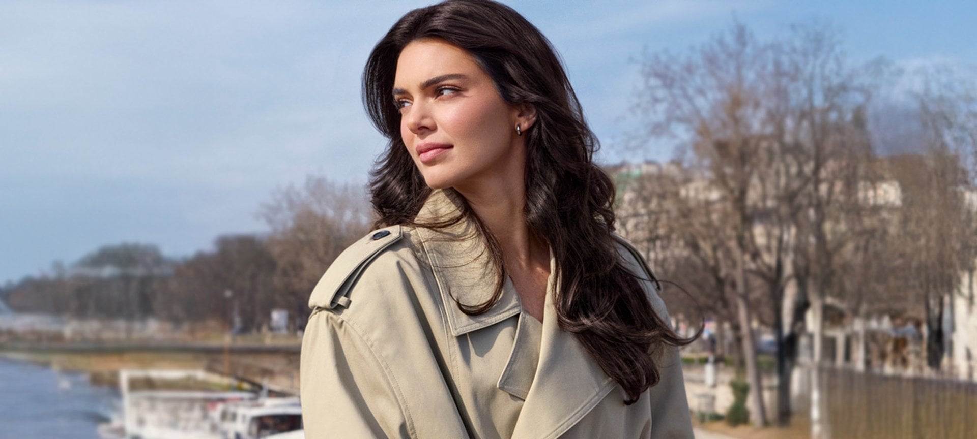 Kendall Jenner, in a three-quarter shot, looks to the right with a slight smile. Her long dark hair is styled in soft waves. She is wearing a beige trench coat and is outdoors, with a blurred background of a bright sky, trees, and what appears to be a body of water or a bridge