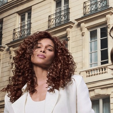 Two women with long, wavy brown hair. The woman on the left has tighter curls and is looking directly at the camera, wearing a white top and a light-colored jacket. The woman on the right has looser waves, looks over her shoulder, and wears a pink top. They are outdoors with a classic European building in the background.