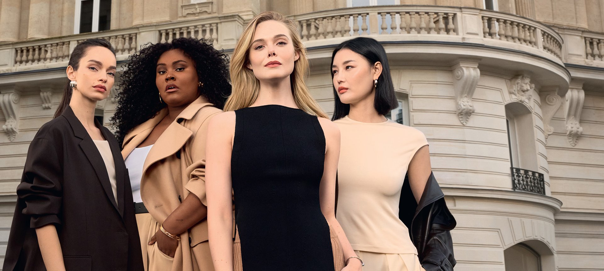A diverse group of four women standing confidently in front of a classic Parisian Haussmann-style building. They are wearing elegant, neutral-toned fashion including a black sleeveless dress, a beige trench coat, and a brown blazer. The image celebrates diversity and sophisticated beauty.