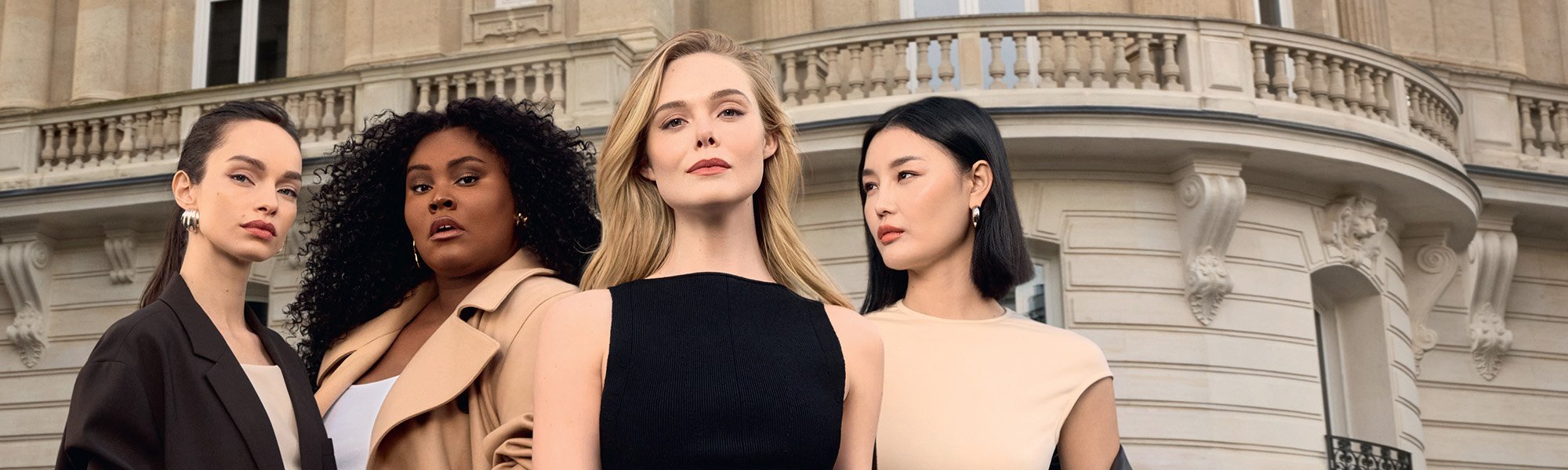 Un groupe de quatre femmes issues de la diversité, posant avec assurance devant un immeuble haussmannien à Paris. Elles portent des tenues élégantes aux tons neutres, incluant une robe noire sans manches, un trench beige et un blazer marron. Cette image célèbre la diversité et la beauté sophistiquée.