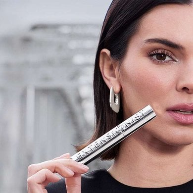 Portrait de l'ambassadrice Kendall Jenner présentant le mascara Telescopic Extensionist de L'Oréal Paris. Elle tient le tube au fini chrome argenté près de son visage, mettant en valeur ses cils longs et parfaitement définis. Elle porte des boucles d'oreilles argentées élégantes sur un fond urbain flou.