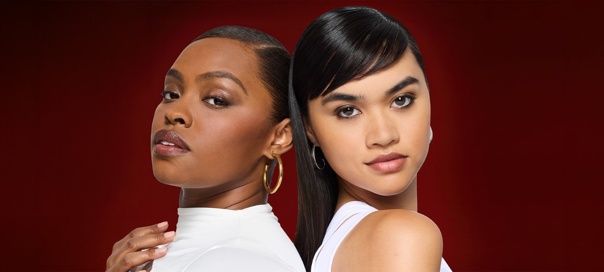 Portrait of two women with different skin tones posing back-to-back against a deep red background. They showcase a flawless, matte complexion, highlighting the inclusivity and efficacy of the Infallible range.