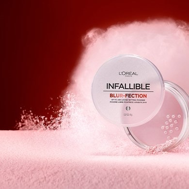 Image artistique présentant le boîtier de poudre libre Infallible Blur-fection. Le poudrier est ouvert au centre d'une explosion de poudre rose pâle soyeuse, illustrant la légèreté et l'effet flouteur du produit sur fond rouge sombre.