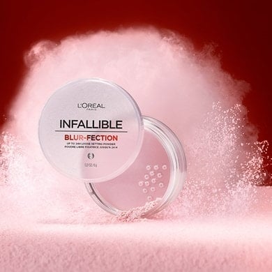 Artistic image featuring the Infallible Blur-fection loose setting powder jar. The compact is open amidst an explosion of silky pale pink powder, showcasing the product's lightness and blurring effect against a dark red background.
