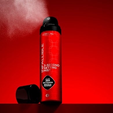 Promotional photography of a metallic red L'Oréal Paris Infallible 3-Second Setting Mist bottle. The spray is active, releasing a cloud of fine mist against a vibrant, dynamic red background.