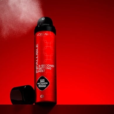 Photographie publicitaire d'un flacon de brume fixatrice Infallible 3-Second Setting Mist de couleur rouge métallique. Le flacon est ouvert et libère un nuage de pulvérisation fine sur un fond rouge vif et dynamique.