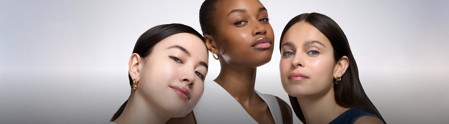 Trois jeunes femmes aux teintes de peau diverses et à la peau éclatante et nette, posant ensemble de près sur un fond clair et simple.