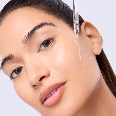 Close-up of a woman with radiant, glowing skin as a drop of liquid is applied to her cheek from a glass dropper.