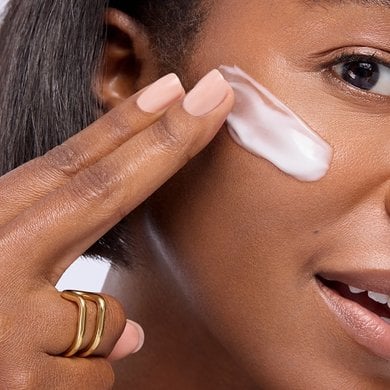 Gros plan d'une femme souriante à la peau mate et au teint éclatant, appliquant une trace de crème blanche ou de soin hydratant sur sa joue avec ses doigts.