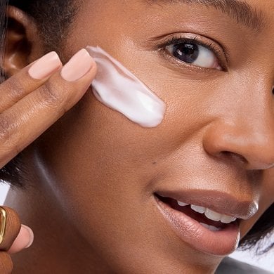 A close-up of a smiling woman with a medium skin tone and radiant, glowing skin applying a streak of white cream or moisturizer to her cheek with her fingers.