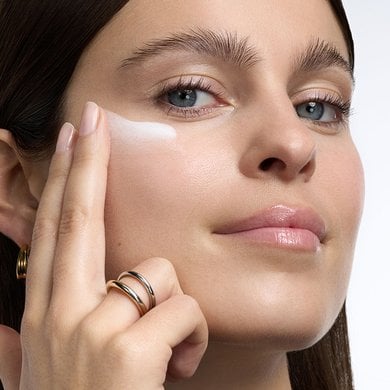 A close-up of a woman with a light skin tone and radiant, glowing skin, applying a streak of white cream or lotion to her cheekbone with her fingers.