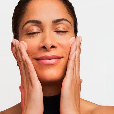 A close-up photograph of a smiling woman with her eyes closed, gently applying a creamy skincare product onto both of her cheeks with her fingertips against a neutral background.