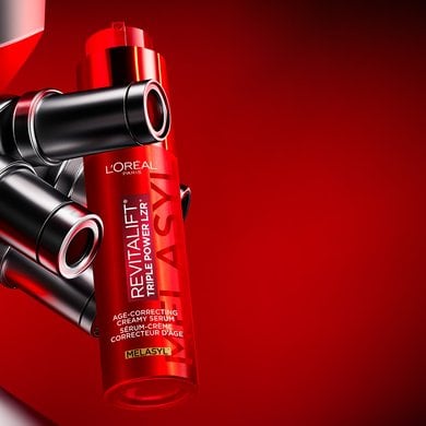 The red L'Oréal Paris Revitalift Triple Power LZR Melasyl serum bottle positioned next to three metallic microscope objective lenses, set against a sleek gradient background shifting from dark red to deep black.
