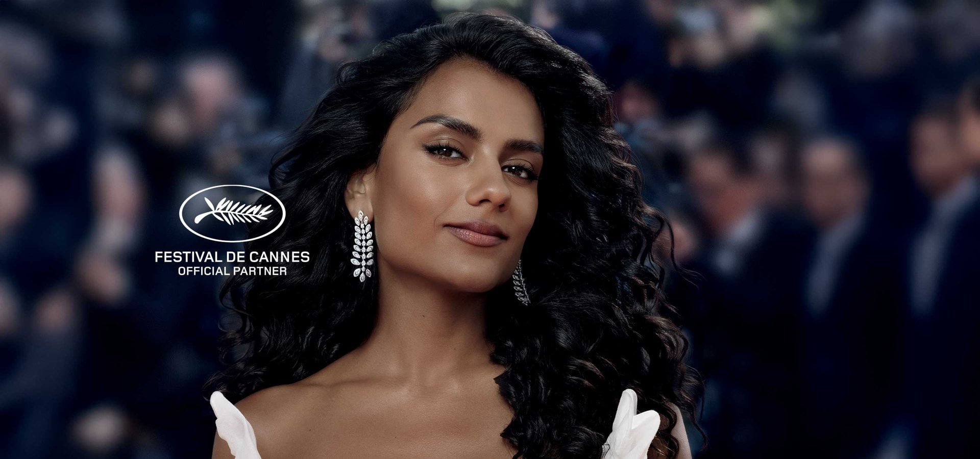 A glamorous portrait of actress Simone Ashley with voluminous wavy dark hair, wearing elegant sparkling leaf-shaped earrings. The background shows a blurred crowd of photographers, and the Festival de Cannes official partner logo is overlaid in the corner.