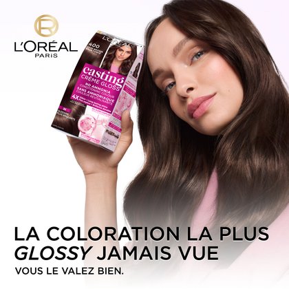 Une femme aux longs cheveux bruns foncés et brillants sourit légèrement, le regard tourné vers le haut. Elle tient une boîte de coloration capillaire L'Oréal Casting Crème Gloss, teinte 400 Brun Foncé, clairement visible avec du texte mettant en avant "SANS AMMONIAQUE," "COULEUR REVITALISANTE," et "plus soyeux + Brillance durable." Le logo L'Oréal Paris est visible dans le coin supérieur gauche. Sous la femme, un grand texte indique "LA COLORATION LA PLUS GLOSSY JAMAIS VUE" et "VOUS LE VALEZ BIEN."