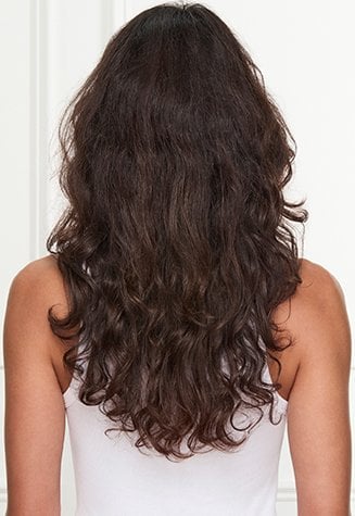 Back view of a woman with long, dark brown hair, styled with more natural waves or texture. She is wearing a white tank top against a pink and white background.