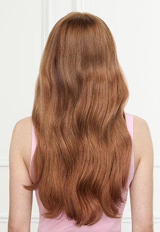 Back view of a woman with long, smooth, shiny coppery red hair with soft waves. She is wearing a pink tank top against a pink and white background.