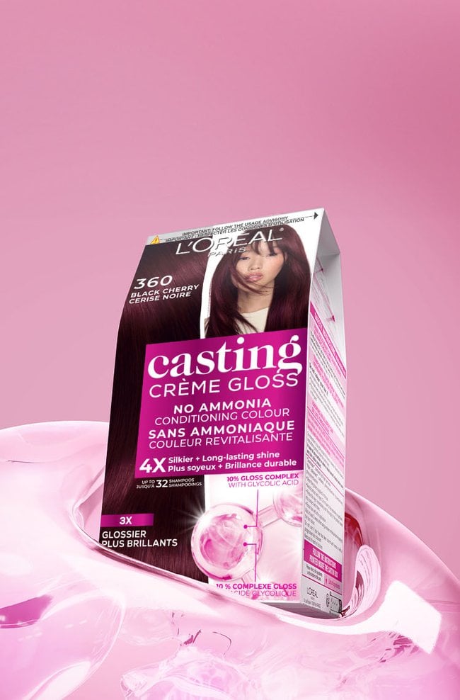 Une boîte noire et rose de coloration capillaire L'Oréal Casting Crème Gloss, nuance 360 Cerise Noire, est dynamiquement posée sur une surface liquide rose brillante et fluide, devant un fond rose en dégradé. La boîte affiche en évidence une femme aux cheveux longs et foncés présentant une riche teinte cerise noire. Les informations clés sur l'emballage mentionnent "SANS AMMONIAQUE COULEUR REVITALISANTE," une promesse de "4X Plus soyeux + Brillance durable" pour "jusqu'à 32 shampooings," et l'intégration d'un "10% COMPLEXE GLOSS AVEC ACIDE GLYCOLIQUE" pour des résultats "3X GLOSSIER PLUS BRILLANTS."