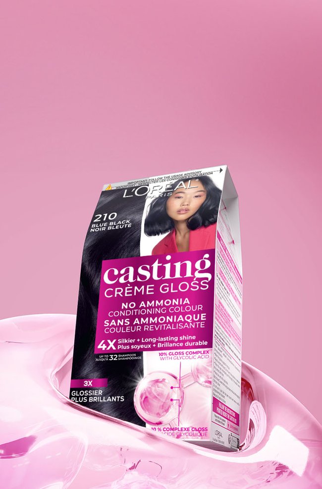 Une boîte noire et rose éclatante de coloration capillaire L'Oréal Casting Crème Gloss, de la nuance 210 Noir Bleuté, est posée sur une surface liquide rose brillante et fluide, devant un fond rose en dégradé. La boîte présente en évidence l'image d'une femme aux cheveux foncés et lisses, avec une subtile nuance bleue. Les principales caractéristiques soulignées sur l'emballage incluent "SANS AMMONIAQUE COULEUR REVITALISANTE," la promesse de "4X Plus soyeux + Brillance durable" pour "jusqu'à 32 shampooings," et la présence d'un "10% COMPLEXE GLOSS AVEC ACIDE GLYCOLIQUE" pour des résultats "3X GLOSSIER PLUS BRILLANTS."