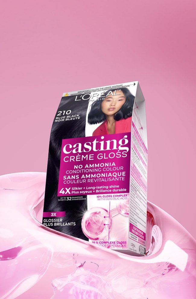 A vibrant black and pink box of L'Oréal Casting Crème Gloss hair color, specifically shade 210 Blue Black, is positioned on a glossy, flowing pink liquid surface against a soft pink gradient background. The box prominently displays an image of a woman with sleek, dark hair that has a subtle blue tint. Key features highlighted on the packaging include "NO AMMONIA CONDITIONING COLOUR," a promise of "4X Silkier + Long-lasting shine" for "up to 32 shampoos," and the presence of a "10% GLOSS COMPLEX WITH GLYCOLIC ACID" for "3X GLOSSIER PLUS BRILLANTS" (3X glossier, more brilliant results).