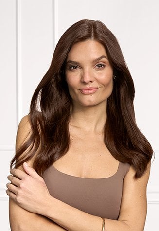 Portrait de la même femme souriante, maintenant avec de longs cheveux châtains foncés, uniformément colorés, sans racines grises visibles. Ses cheveux apparaissent brillants et sains, sur un fond clair.