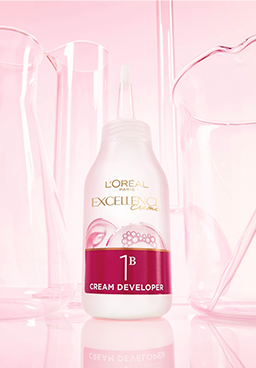 Product photography showing a white bottle of L'Oréal Paris Excellence Crème developer cream (step 1B). The bottle is centered and surrounded by artistic, transparent laboratory glassware against a soft, bright pastel pink background, evoking scientific research and product formulation.