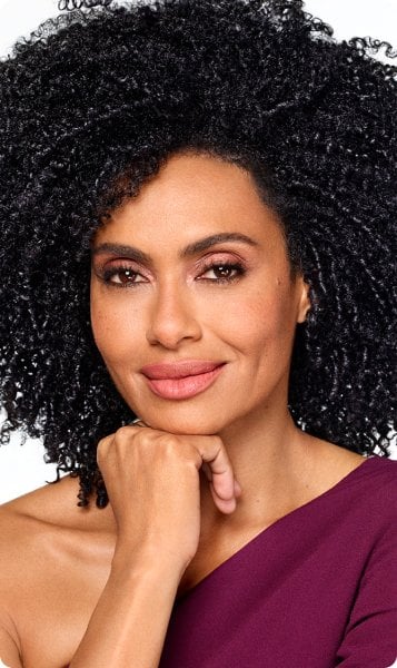 Portrait of a woman with voluminous, tightly curled black hair. She is looking at the camera with a slight smile, her hand resting under her chin. She is wearing a plum-colored top.
