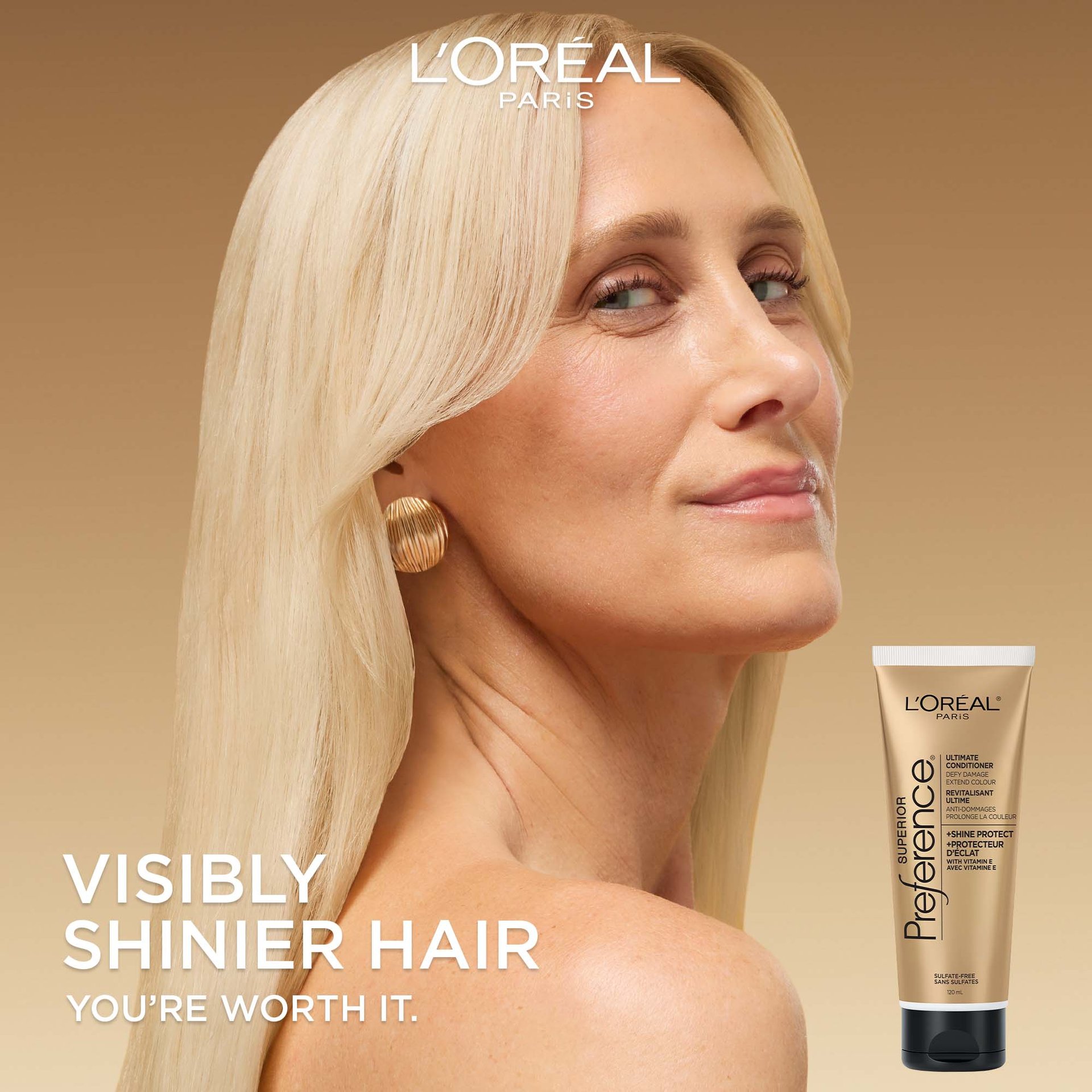A woman with sleek, long blonde hair looks over her shoulder with a slight smile. The background is a soft gold gradient. Large white text on the left reads "VISIBLY SHINIER HAIR. YOU’RE WORTH IT." An image of the conditioner tube is in the bottom right corner.