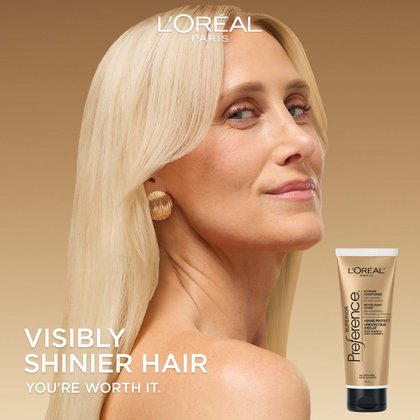 A woman with sleek, long blonde hair looks over her shoulder with a slight smile. The background is a soft gold gradient. Large white text on the left reads "VISIBLY SHINIER HAIR. YOU’RE WORTH IT." An image of the conditioner tube is in the bottom right corner.